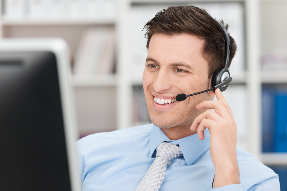 Smiling friendly handsome young male call centre operator or client services personnel beaming as he listens to a call and checks information on his computer monitor